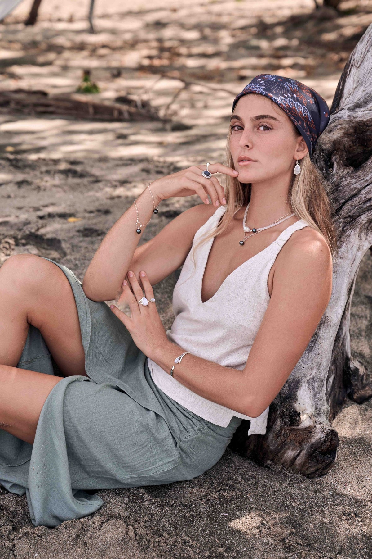 A woman sitting outdoors against a large tree trunk, wearing a white sleeveless top, sage green skirt, and a blue patterned headscarf. She is styled with jewelry including a silver necklace, bracelet, and rings.
