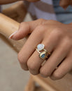 May include: Two gold rings with gemstones. One ring features a round, white gemstone, while the other has a teardrop-shaped, blue-green gemstone. The rings are worn together on a finger, showcasing a layered jewelry style. The rings have a textured band.