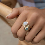 May include: Two gold rings with gemstones. One ring features a round, white gemstone, while the other has a teardrop-shaped, blue-green gemstone. The rings are worn together on a finger, showcasing a layered jewelry style. The rings have a textured band.