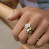 May include: Two gold rings with gemstones. One ring features a round, white gemstone, while the other has a teardrop-shaped, blue-green gemstone. The rings are worn together on a finger, showcasing a layered jewelry style. The rings have a textured band.