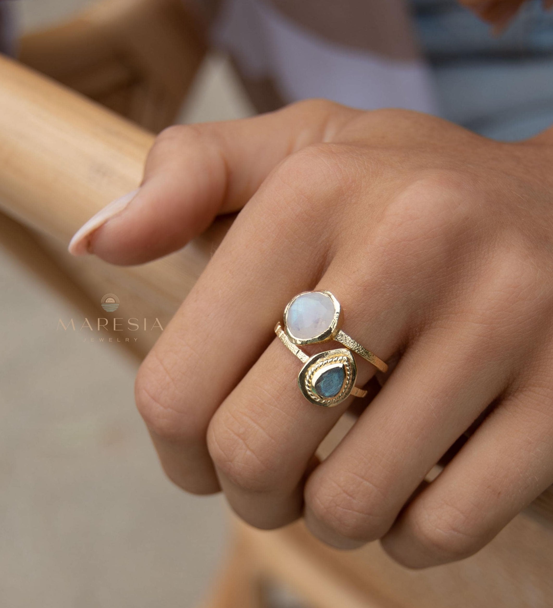 May include: Two gold rings with gemstones. One ring features a round, white gemstone, while the other has a teardrop-shaped, blue-green gemstone. The rings are worn together on a finger, showcasing a layered jewelry style. The rings have a textured band.