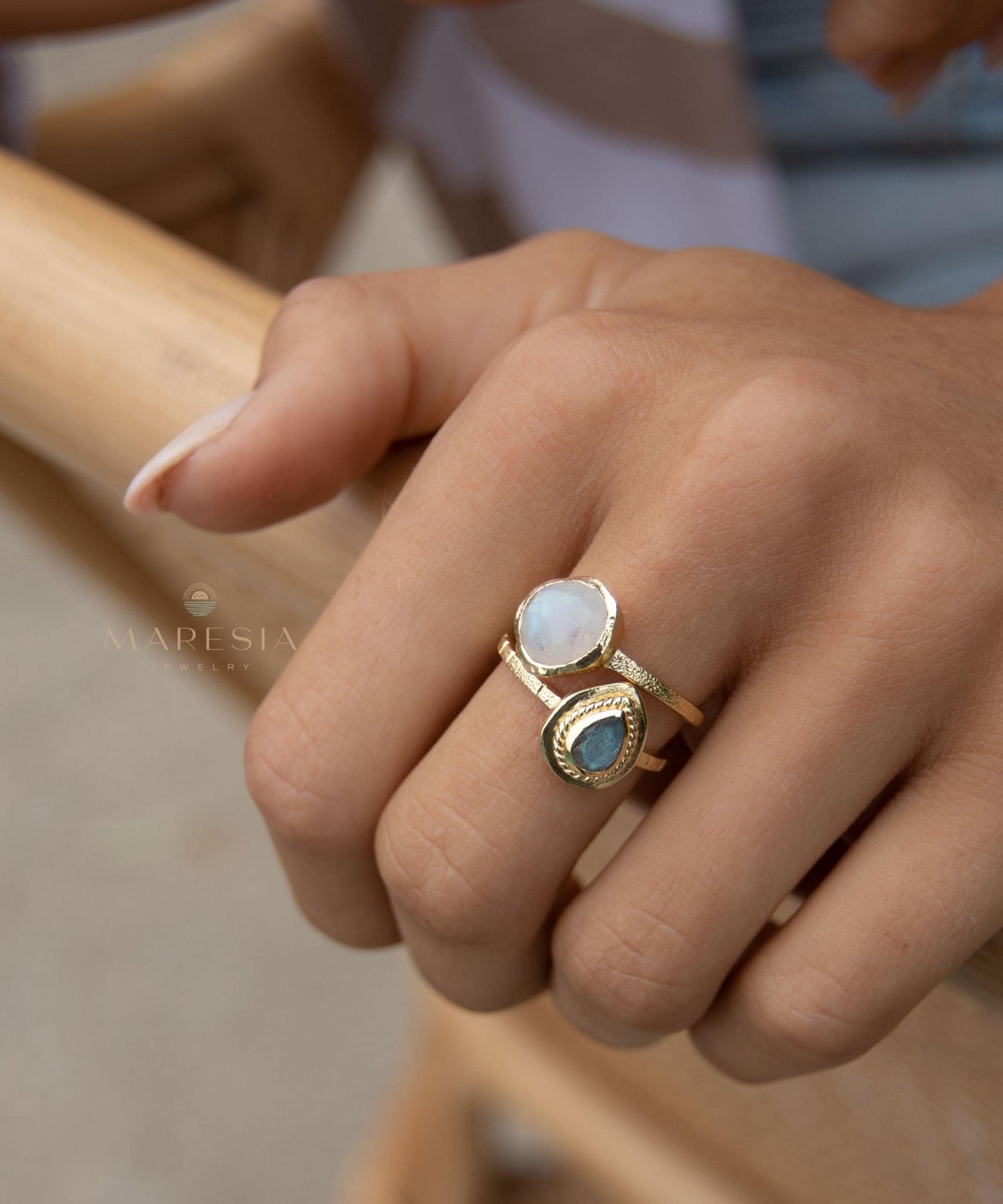 May include: Two gold rings with gemstones. One ring features a round, white gemstone, while the other has a teardrop-shaped, blue-green gemstone. The rings are worn together on a finger, showcasing a layered jewelry style. The rings have a textured band.
