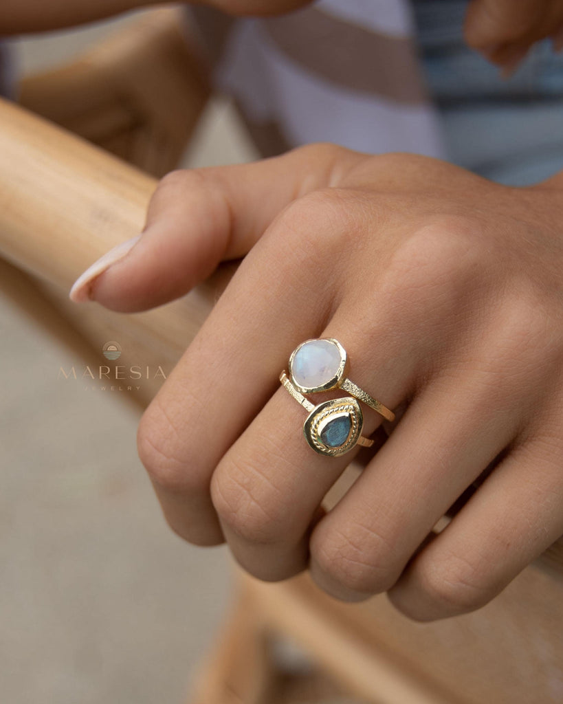 May include: Two gold rings with gemstones. One ring features a round, white gemstone, while the other has a teardrop-shaped, blue-green gemstone. The rings are worn together on a finger, showcasing a layered jewelry style. The rings have a textured band.