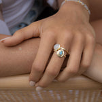May include: Two gold rings with gemstone accents. One ring features a light blue, teardrop-shaped stone, while the other has a white, oval-shaped stone. A delicate gold bracelet with small, round beads is also visible. The rings and bracelet are displayed on a hand.