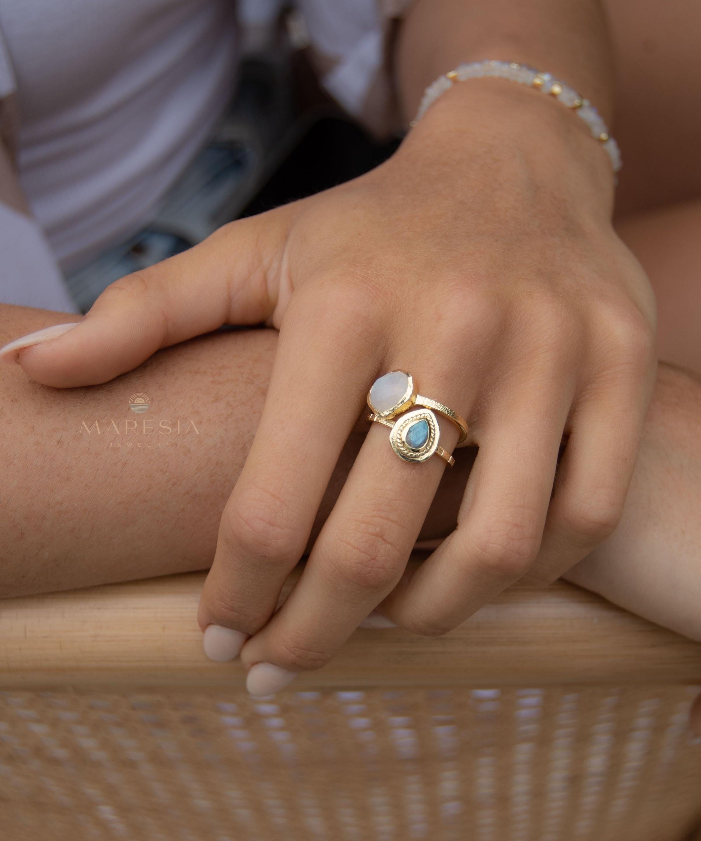 May include: Two gold rings with gemstone accents. One ring features a light blue, teardrop-shaped stone, while the other has a white, oval-shaped stone. A delicate gold bracelet with small, round beads is also visible. The rings and bracelet are displayed on a hand.
