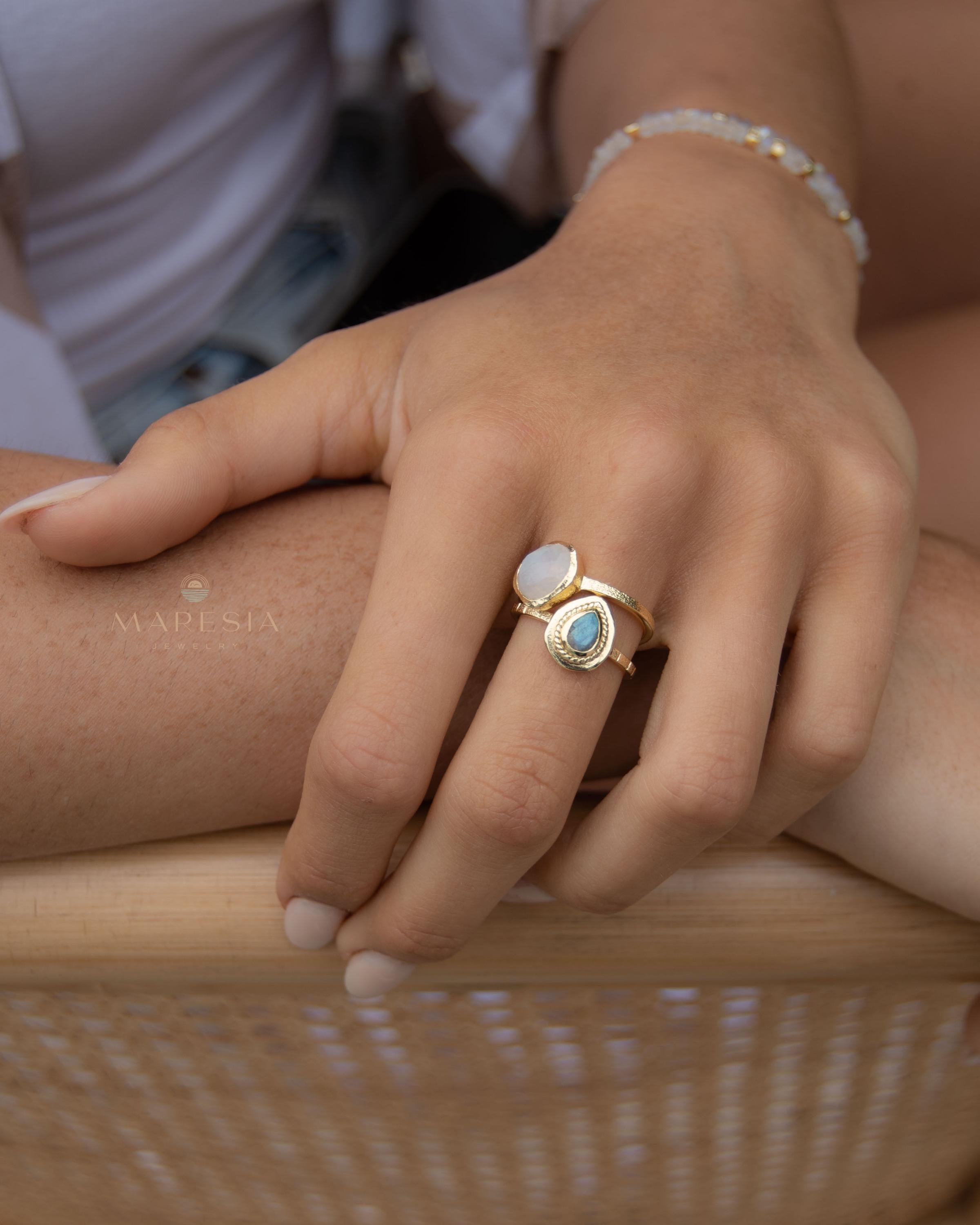 May include: Two gold rings with gemstone accents. One ring features a light blue, teardrop-shaped stone, while the other has a white, oval-shaped stone. A delicate gold bracelet with small, round beads is also visible. The rings and bracelet are displayed on a hand.